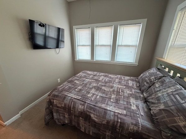 A bedroom with a bed, a television mounted on the wall, and a window with blinds. - 2 Bedroom, main floor room with private entrance (Fairbury)
