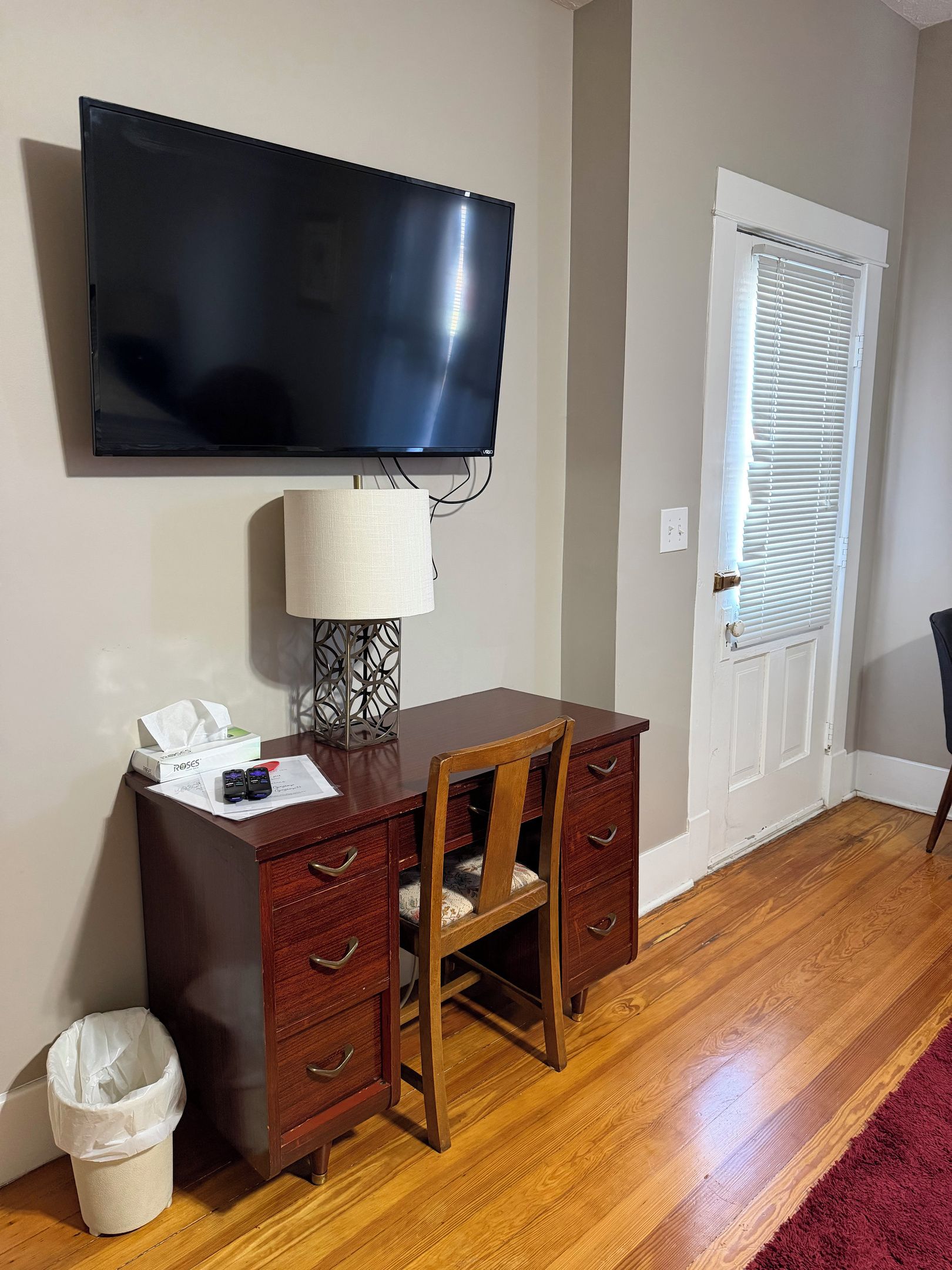 Une pièce dotée d’un téléviseur à écran plat fixé au mur, d’un bureau en bois avec des tiroirs, d’une chaise, d’une lampe et d’une porte avec des stores.