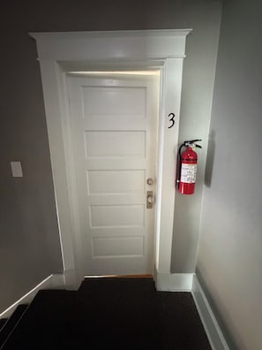 Una porta blanca amb el número 3 a la paret al seu costat, un extintor vermell fixat a la pared, i un sòl amb moqueta.