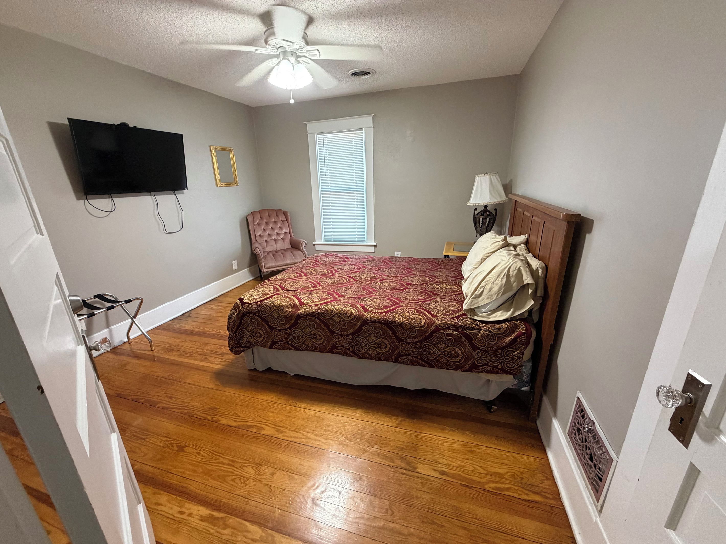 A bedroom with a bed, a ceiling fan, a window, a chair, and a television mounted on the wall.