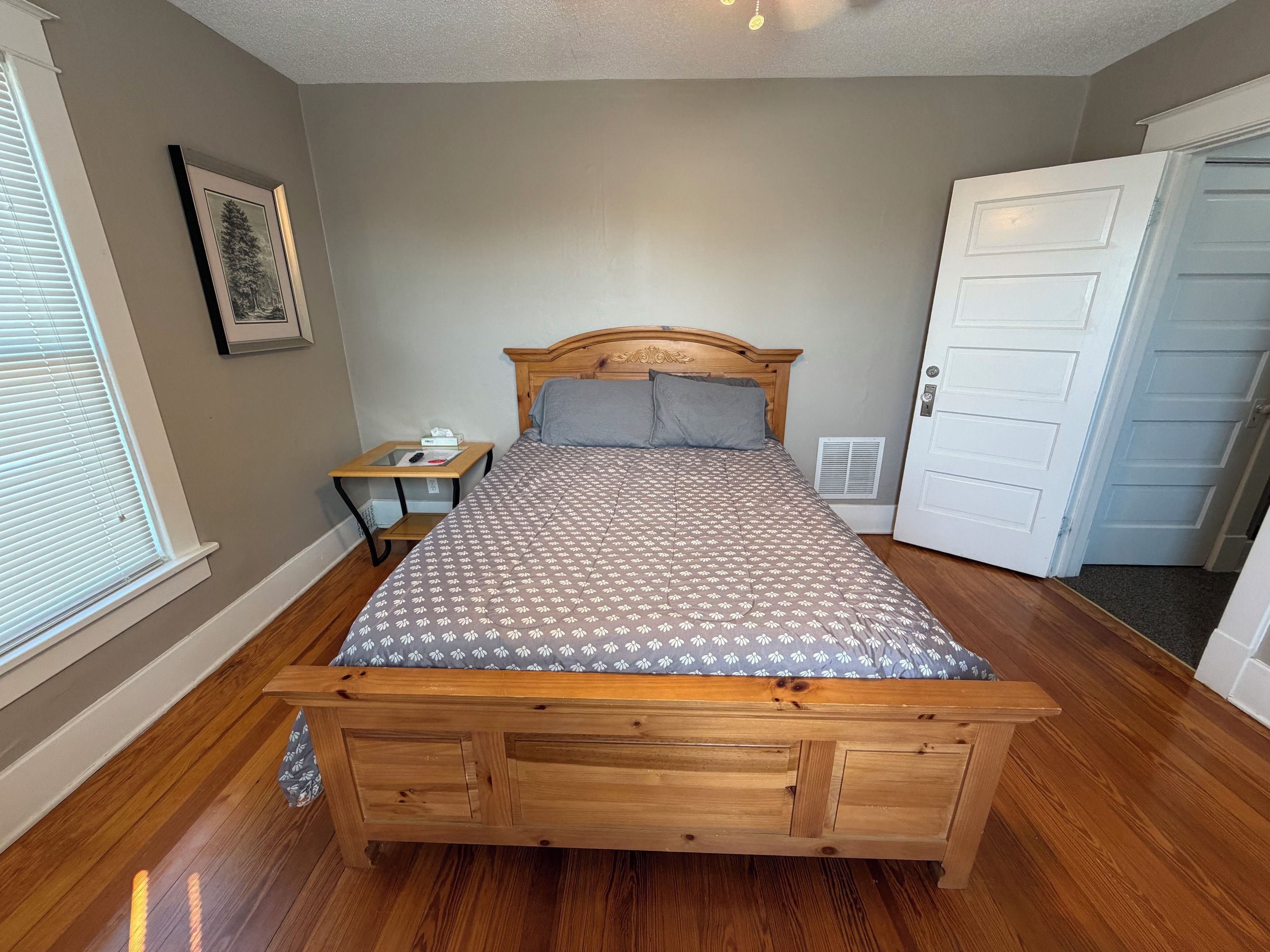 Une chambre à coucher avec un lit en bois, une table de chevet, une commode, un tableau encadré au mur et une porte.