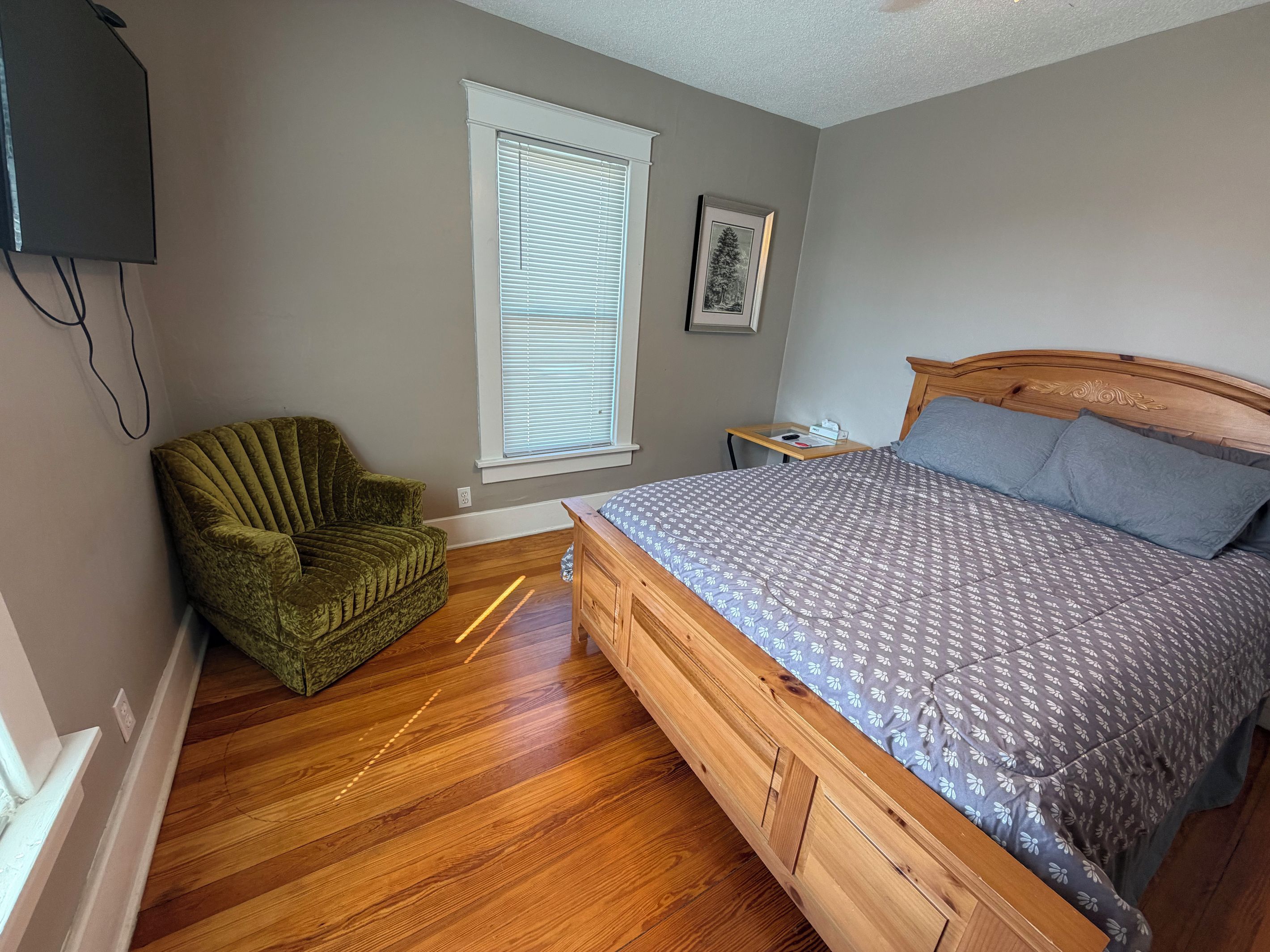 Une chambre à coucher avec un lit en bois, un fauteuil vert, une fenêtre avec des stores et une petite table de chevet.