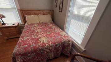 A bedroom with a wooden headboard, floral bedspread, a nightstand with a lamp, and a window with blinds.
