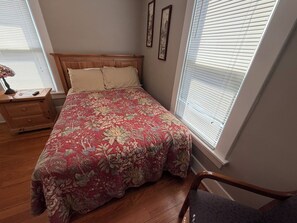 A bedroom with a wooden headboard, floral bedspread, a nightstand with a lamp, and a window with blinds. - 1 Bedroom Unit with Full Size Bed (Fairbury)