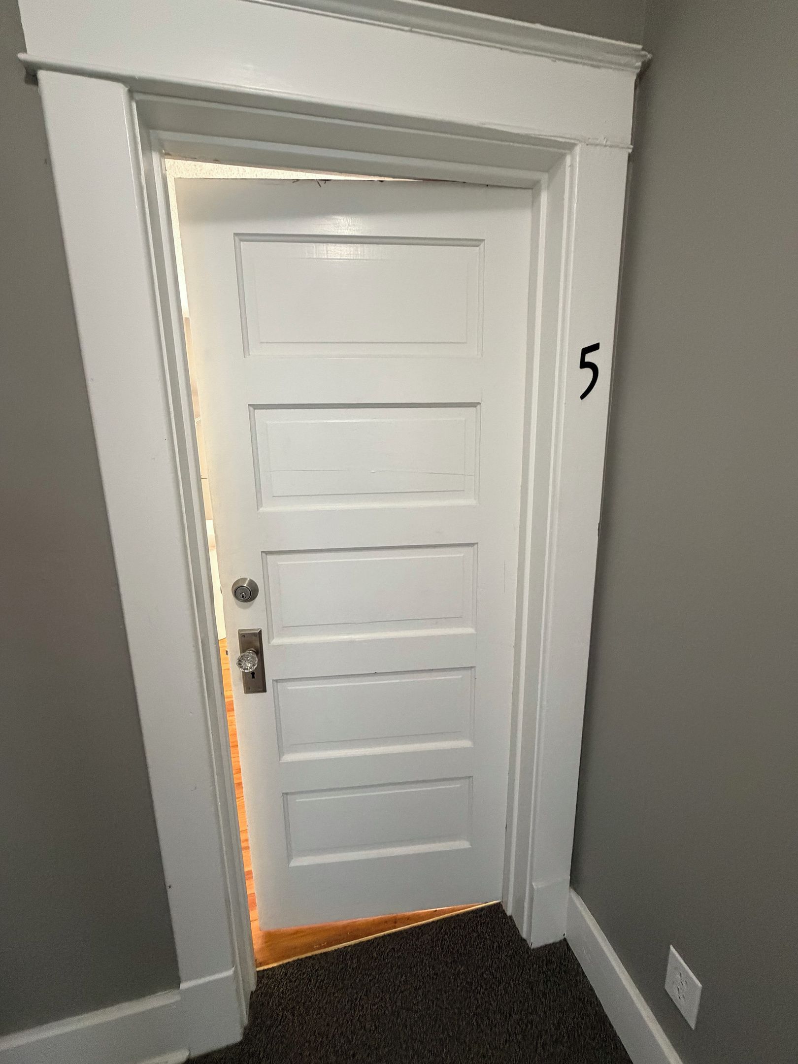 Une porte blanche, avec le chiffre « 5 » peint sur le mur à côté, une poignée de porte et un sol recouvert d’une moquette.