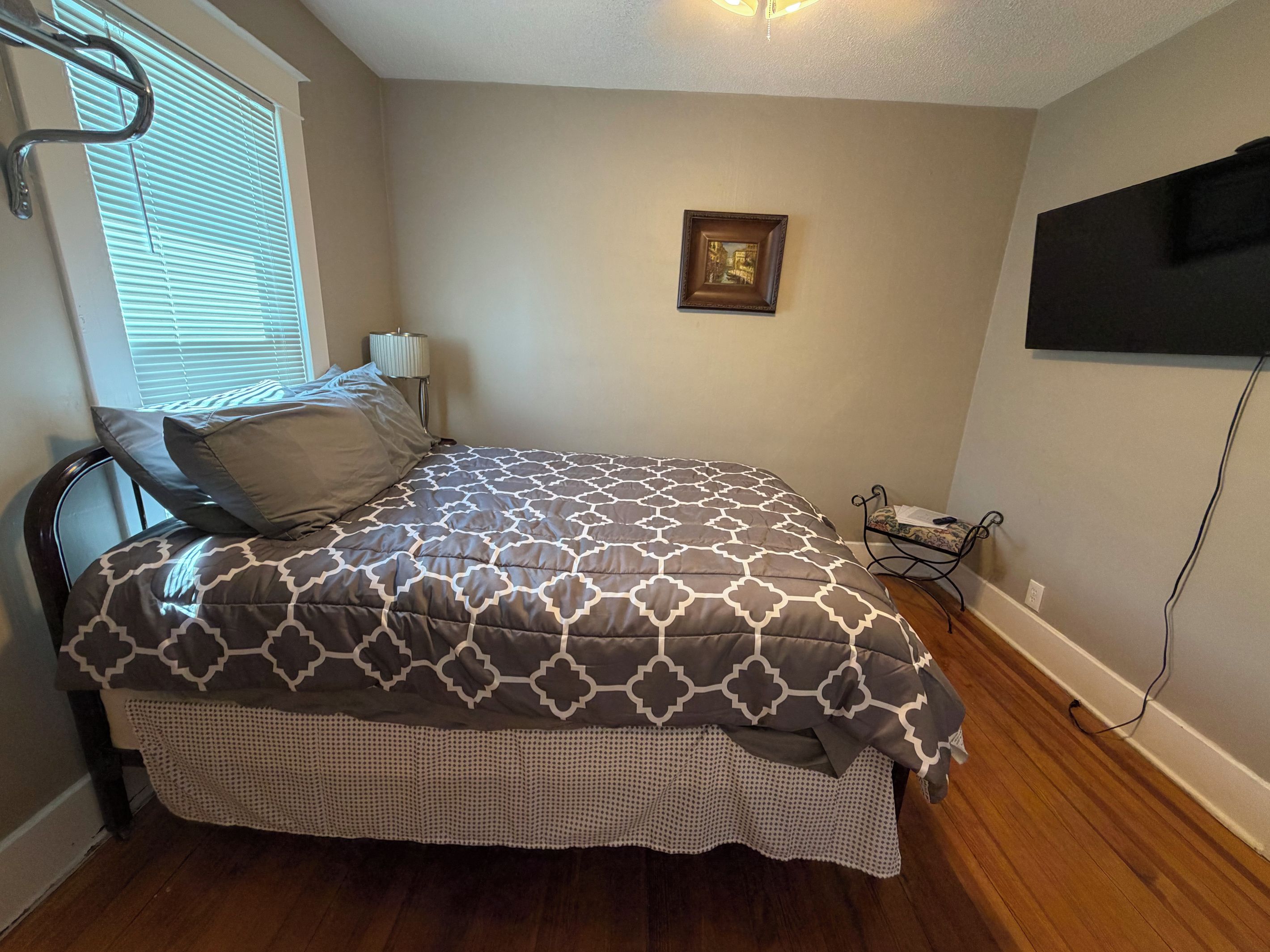 A bedroom with a bed, a window with blinds, a wall-mounted TV, and a bedside table with a lamp.