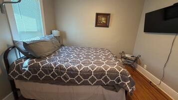 A bedroom with a bed, a window with blinds, a wall-mounted TV, and a bedside table with a lamp.