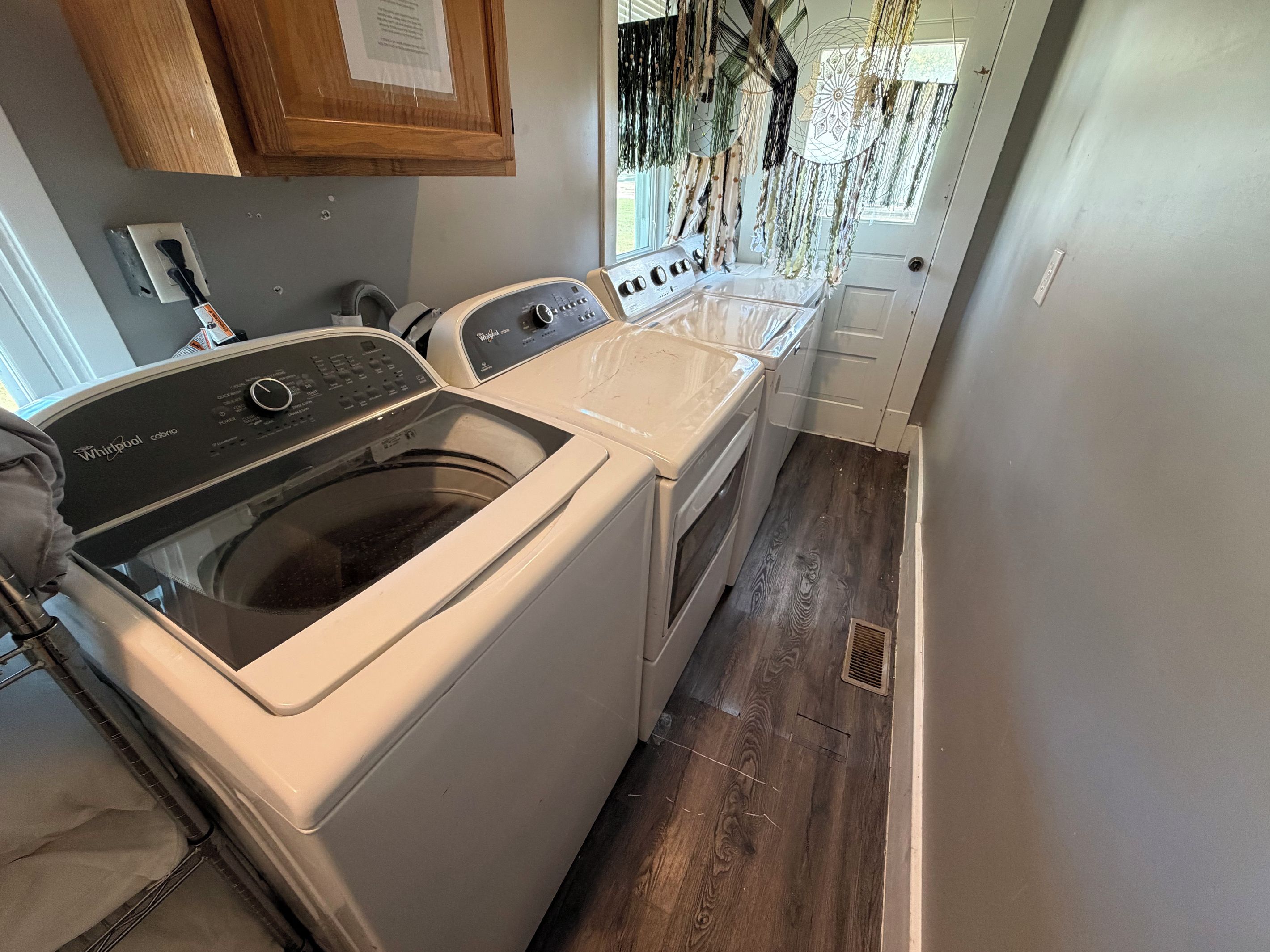 A laundry room with a front-loading washer and dryer, wooden cabinets, and a window with hanging dreamcatchers.