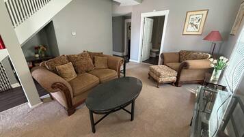 Un salon comprenant un canapé, un pouf et une table basse en verre. On y trouve un escalier avec une rampe, une porte donnant sur une autre pièce et un tableau encadré accroché au mur.