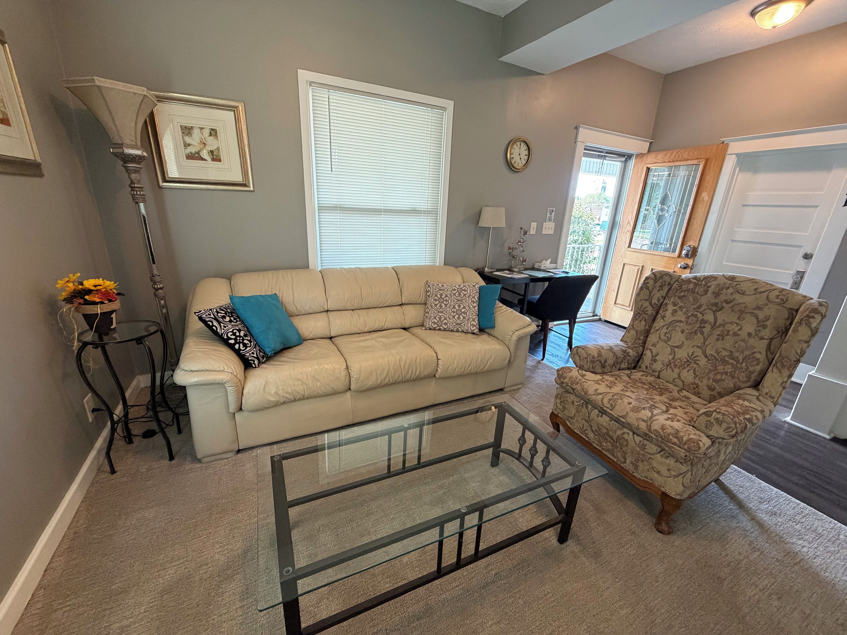 A living room with a beige sofa, a patterned armchair, a glass coffee table, a floor lamp, and a side table with a vase of flowers.