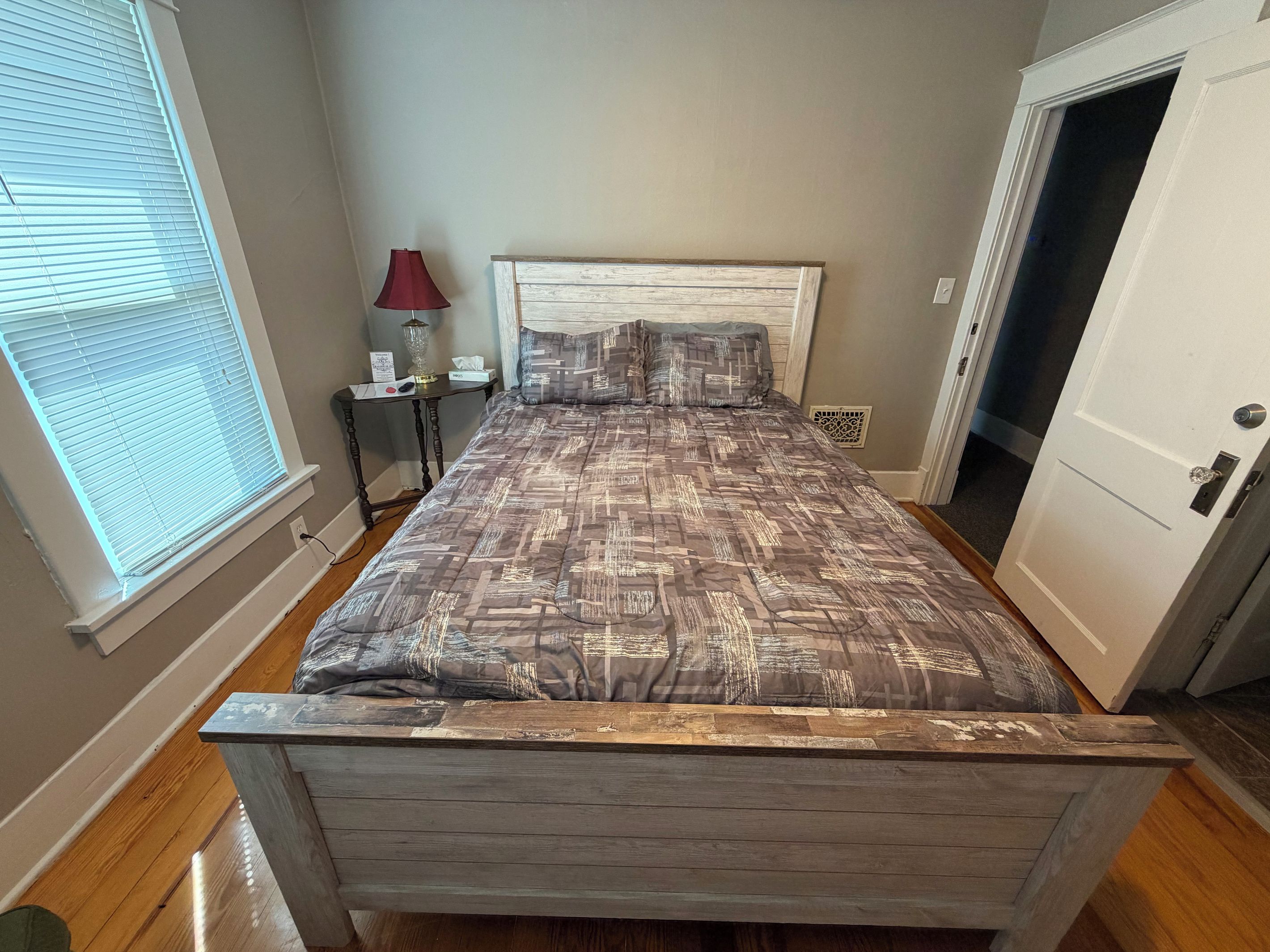 A bedroom with a wooden headboard, a patterned bedspread, a nightstand with a lamp, and a door to another room.