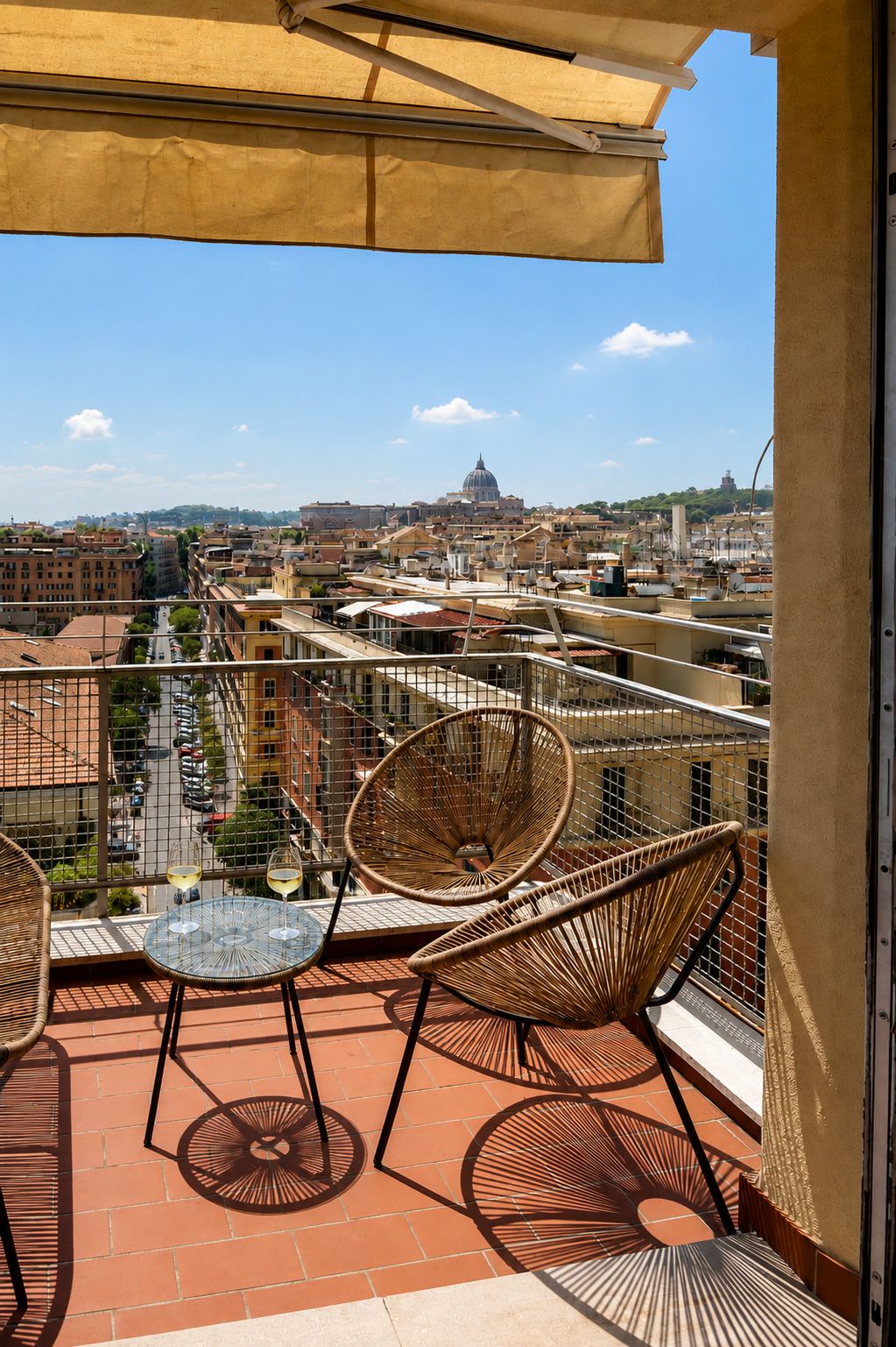A balcony with wicker chairs and a table, overlooking a cityscape with buildings and a dome structure.
