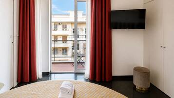 A hotel room with a bed, a flat-screen TV, a round table with a towel, a round ottoman, and a balcony with red curtains.