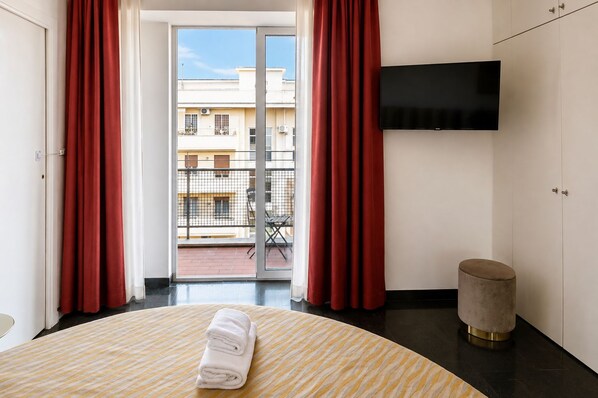 A hotel room with a bed, a flat-screen TV, a round table with a towel, a round ottoman, and a balcony with red curtains. - Vatican Sky view Spa Apartment by RHR (ROMA)