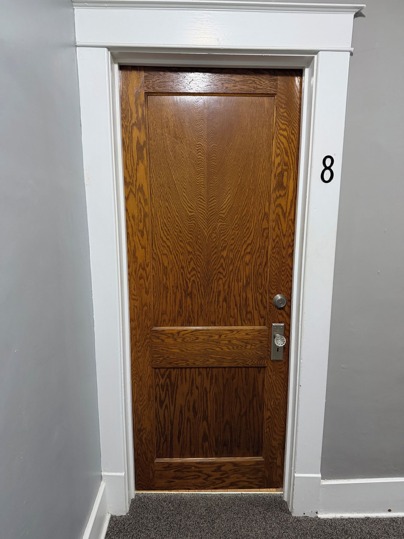 Una puerta de madera con un ojo de cerradura y un cerrojo, el número 8 en la pared, y un suelo alfombrado.