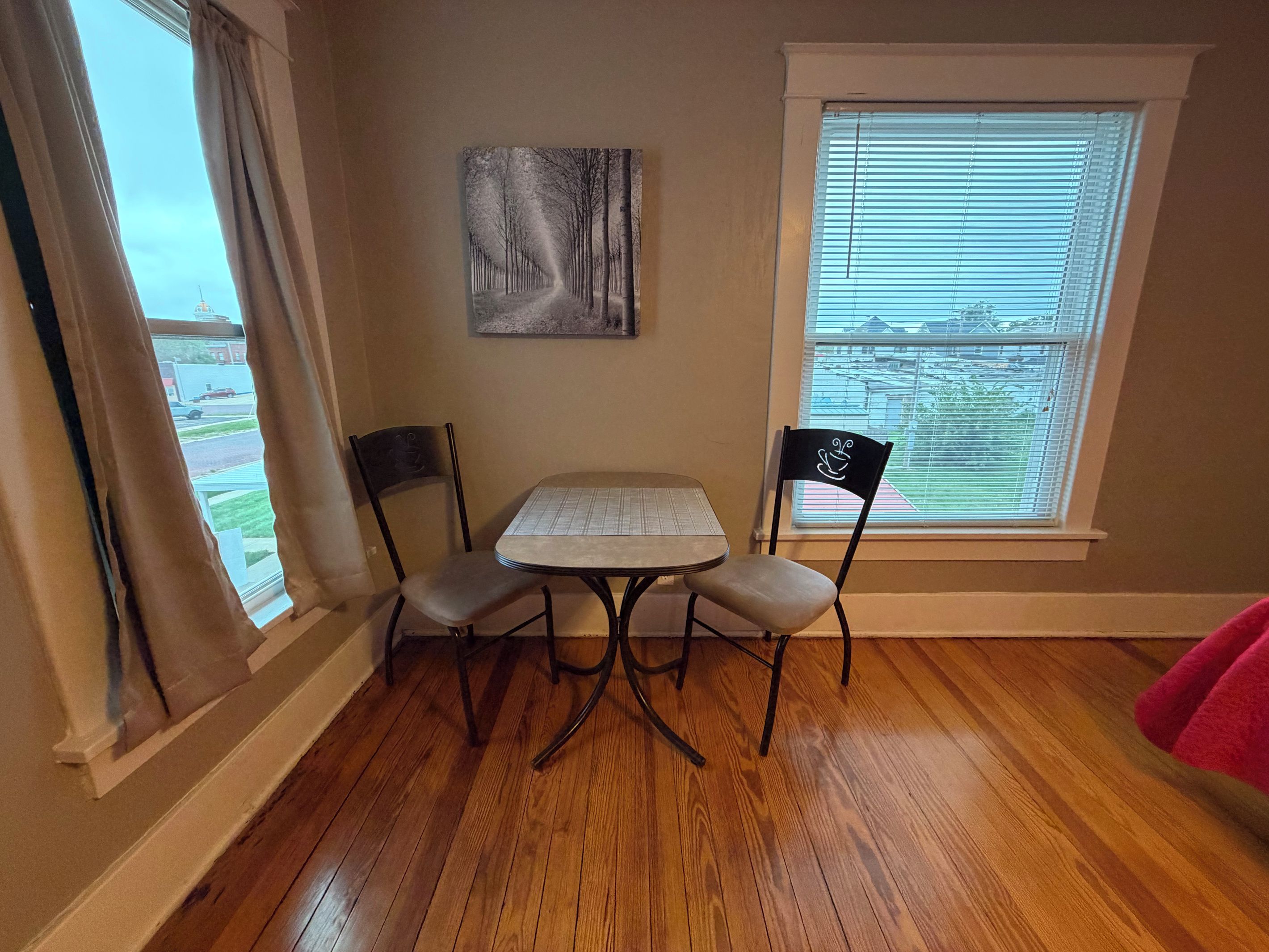 A room with a wooden floor, a table with four chairs, a painting on the wall, and a window with blinds.