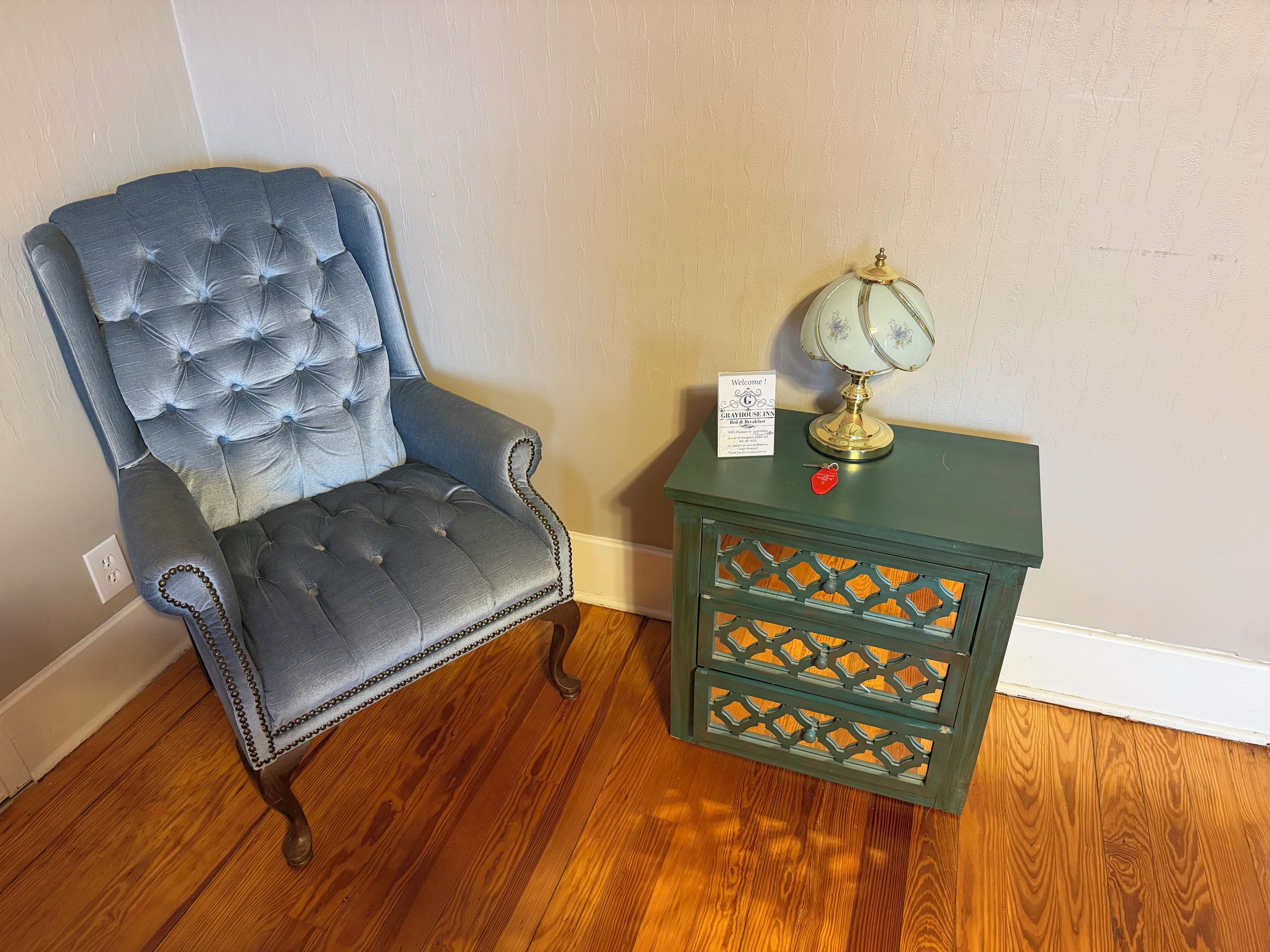 A tufted armchair with a decorative lamp and a sign on a wooden nightstand.