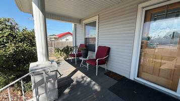 A covered porch with two red chairs and a small table, a wooden deck, and a view of a neighboring house.