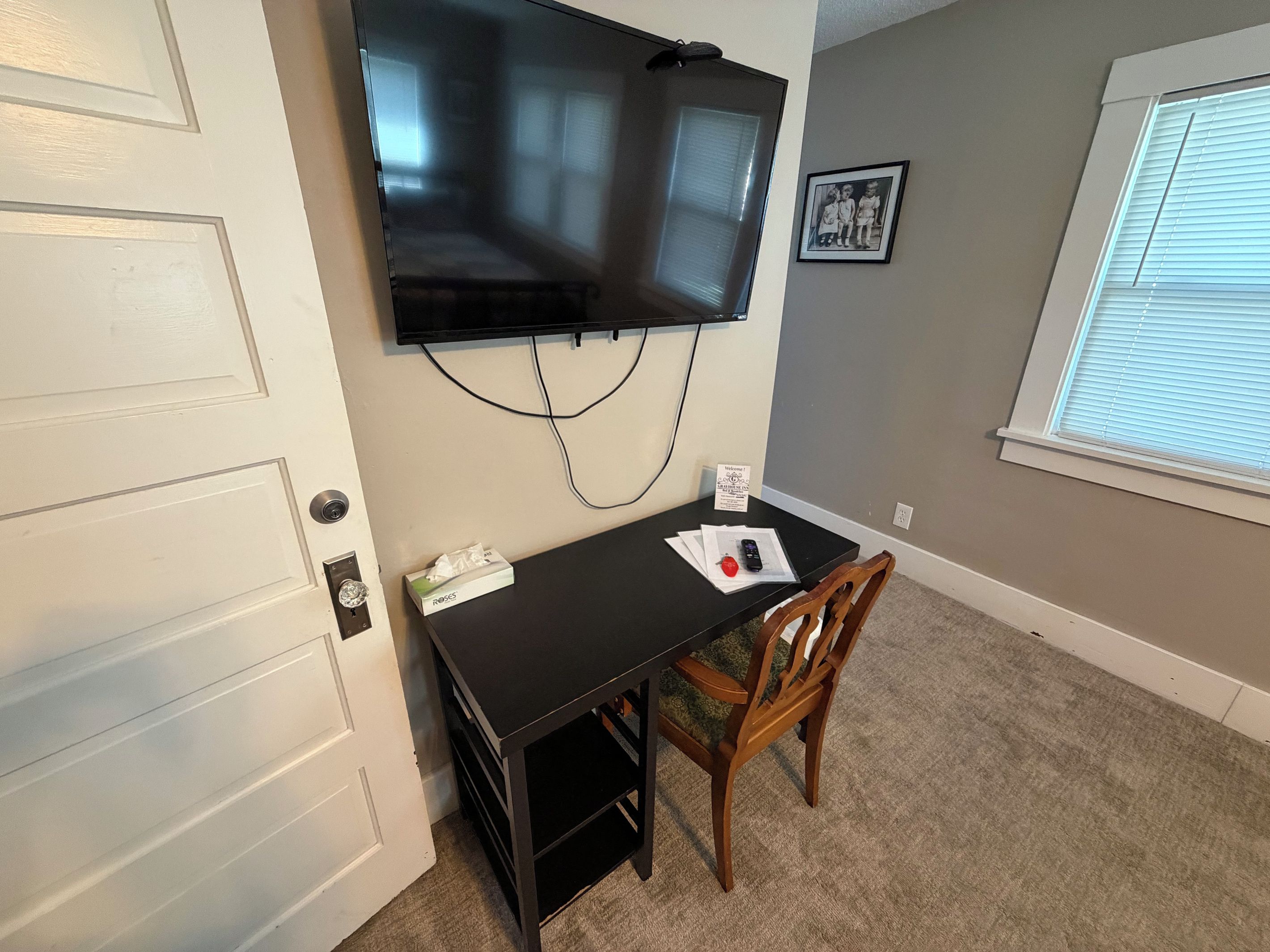Quarto com televisão de tela plana na parede, uma mesa pequena com cadeira e janela com persianas.