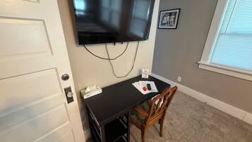 Quarto com televisão de tela plana na parede, uma mesa pequena com cadeira e janela com persianas.