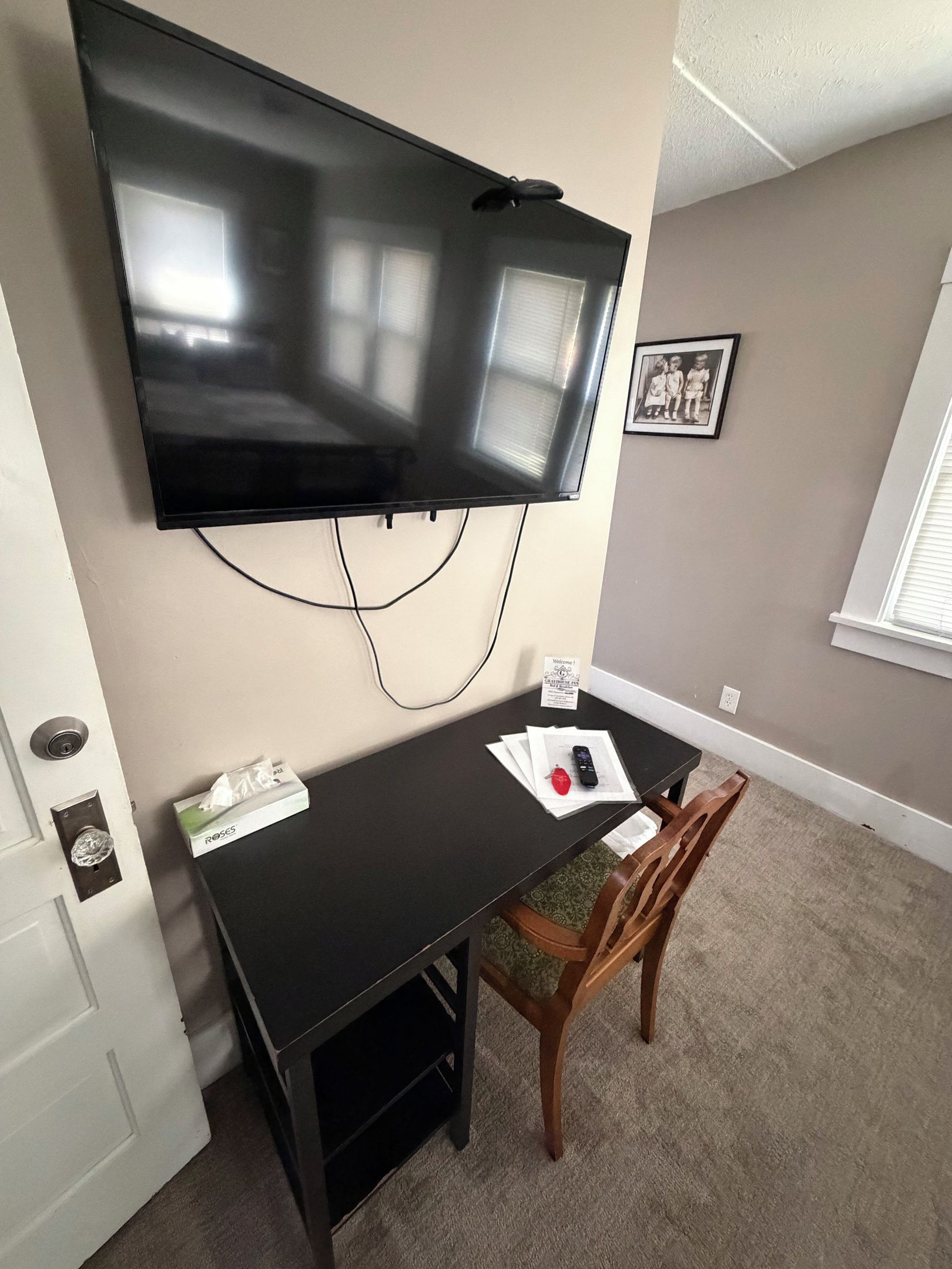 A room with a flat-screen TV mounted on the wall, a small table with a chair, a door with a keyhole, and a framed photo on the wall.