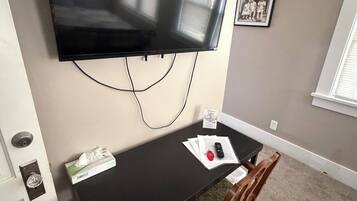 A room with a flat-screen TV mounted on the wall, a small table with a chair, a door with a keyhole, and a framed photo on the wall.