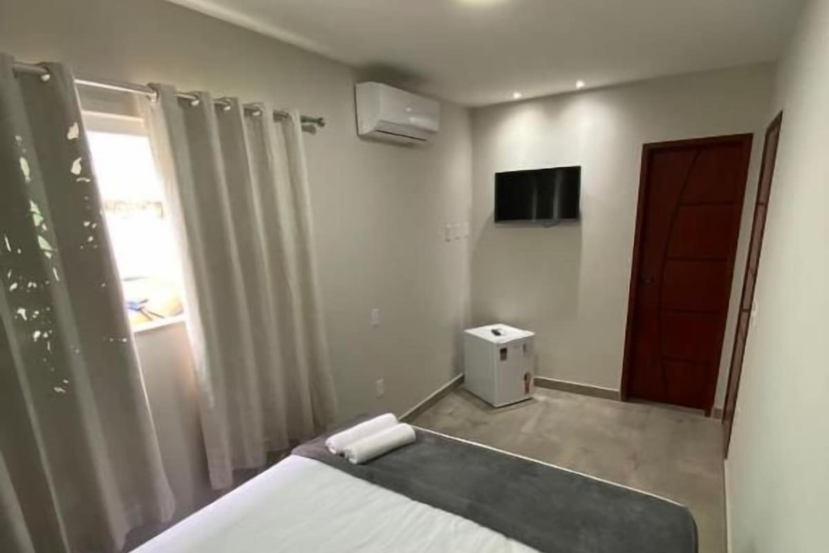 Quarto de hotel com cama, mesa de cabeceira, aparelho de ar condicionado, televisão e porta.