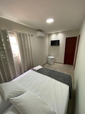 Quarto de hotel com cama, mesa de cabeceira, aparelho de ar condicionado, televisão e porta.