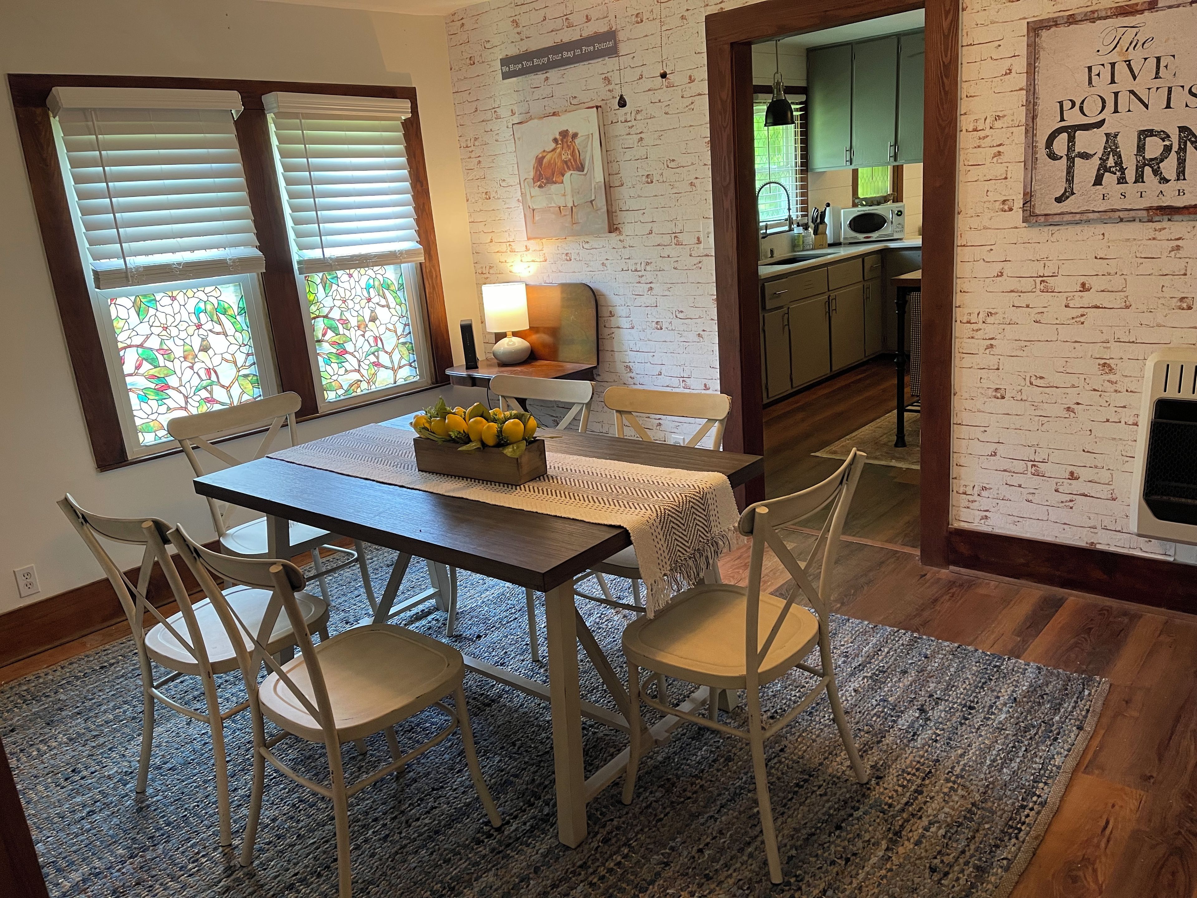 A dining area with a table, chairs, and a fruit centerpiece. There are windows with blinds and a kitchen visible in the background.