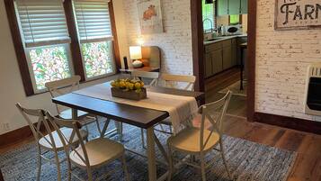 A dining area with a table, chairs, and a fruit centerpiece. There are windows with blinds and a kitchen visible in the background.