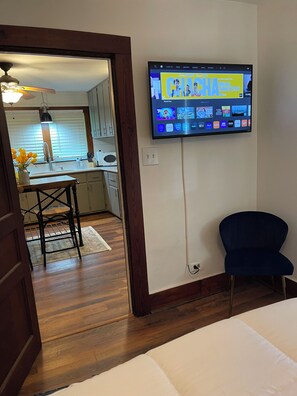 Een kamer met een bed, een aan de muur bevestigde televisie waarop diverse streamingdiensten worden getoond, een donkerblauwe stoel en een houten vloer.