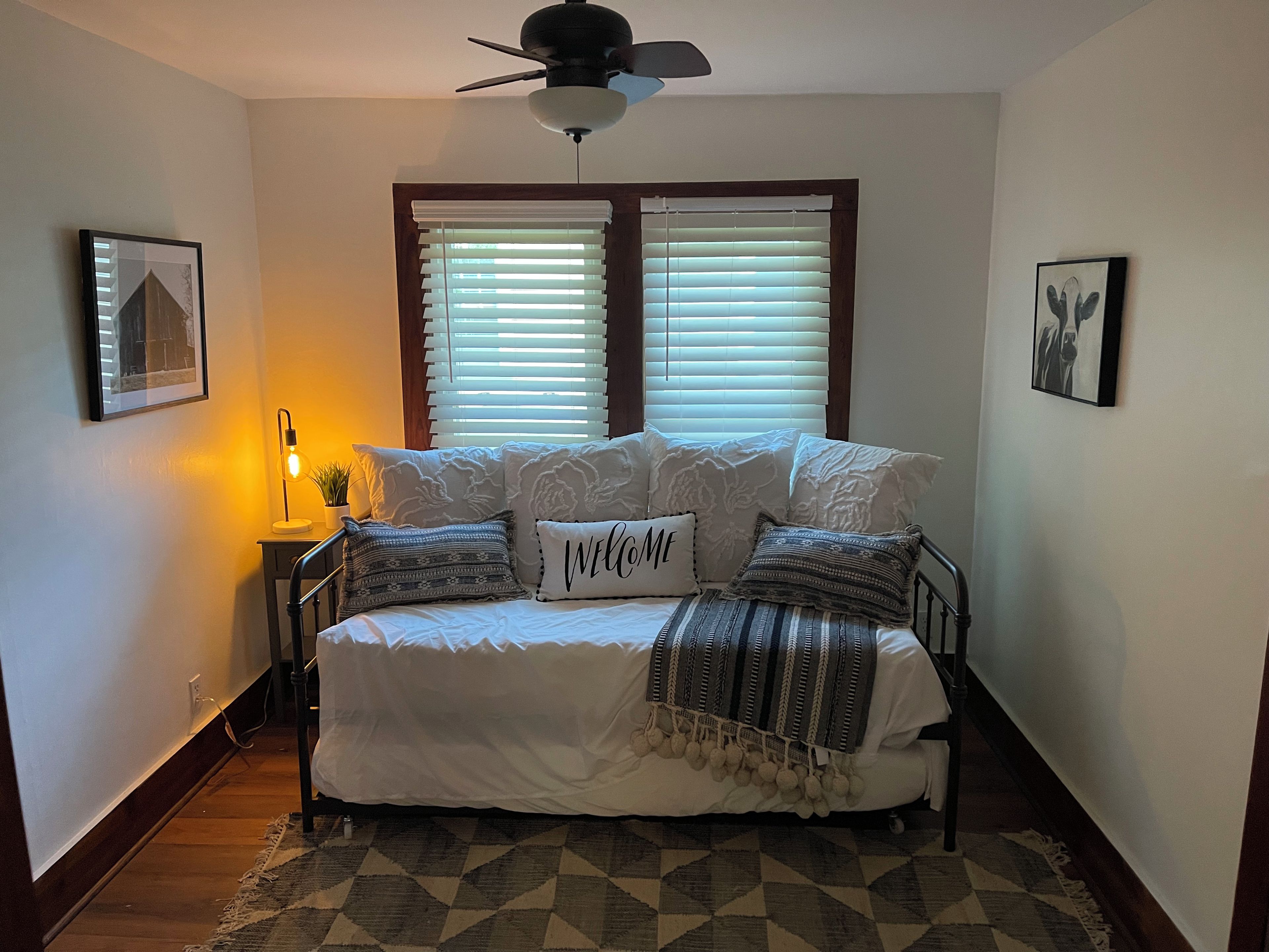 Un dormitorio con cama, ventilador de techo, ventana con persianas, lámpara de noche y alfombra.