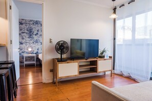 Un salon moderne avec un téléviseur à écran plat posé sur une console en bois, un ventilateur sur pied et une petite table avec une lampe.