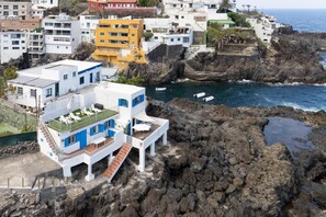 Um edifício costeiro com terraço no telhado, próximo a terreno rochoso e um corpo de água.
