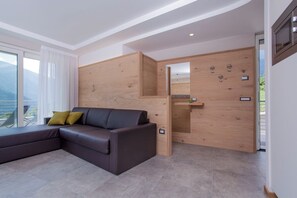 A modern living room with a sectional sofa, wooden panel walls, a small wooden shelf, and a view through a large window. - CAMELIE APARTMENT - BELLAVISTA LAKE RESIDENCE (Trentino-Alto Adige)