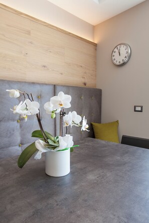 Quarto moderno com mesa de concreto cinza, um vaso branco com flores brancas, cabeceira estofada cinza, almofada amarelo mostarda e um relógio de parede.