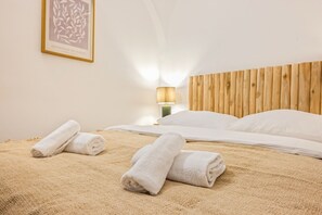 Une chambre d’hôtel avec une tête de lit en bois, un lit recouvert de draps blancs, deux serviettes enroulées posées sur le lit et une lampe fixée au mur.