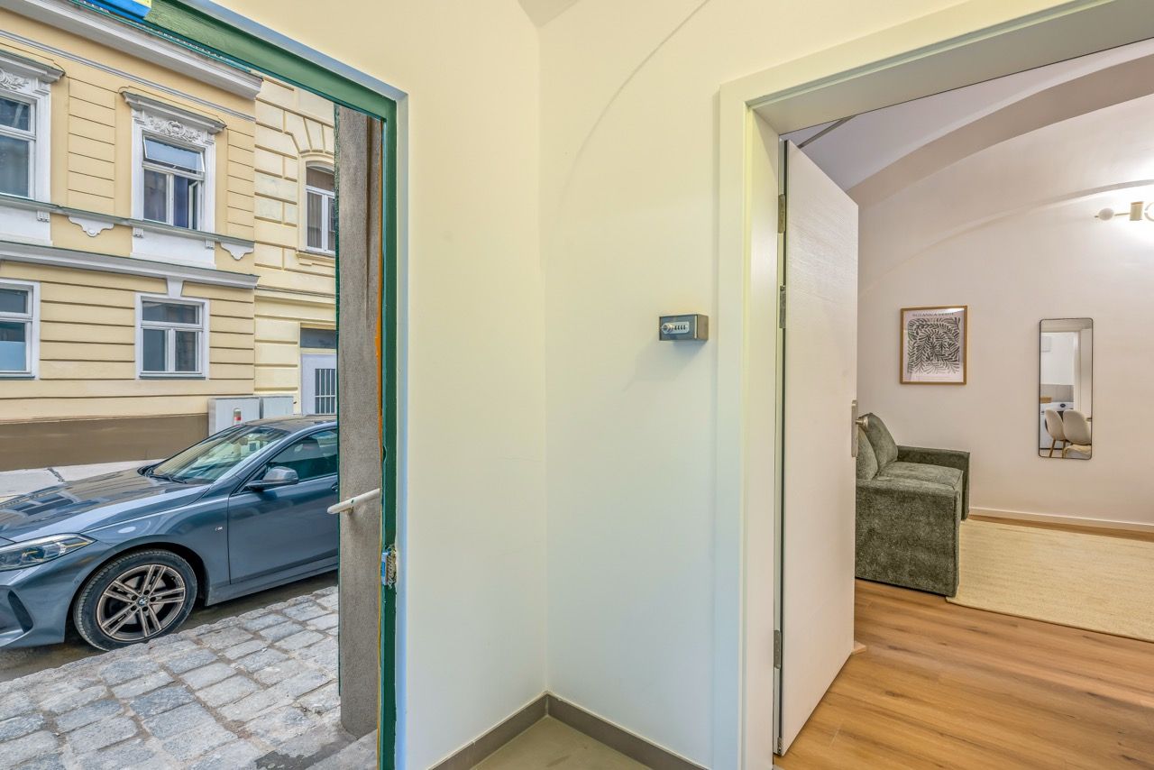 Ein Flur mit einem geparkten Auto vor der Tür, einer Tür, die zu einem Zimmer mit einem Sessel und einem Spiegel führt, und einem Gebäude mit Fenstern im Hintergrund.