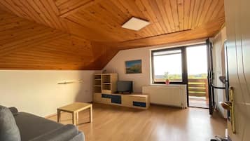 Una habitación con techo de madera inclinado, un sofá, un mueble para televisión, un radiador y una ventana con vistas.