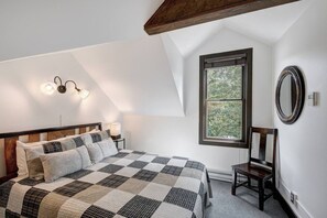 Ein Schlafzimmer mit karierter Bettdecke, einem hölzernen Kopfteil, einem Nachttisch mit Lampe, einem Fenster mit Blick auf Bäume und einem Wandspiegel.