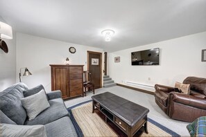 Een woonkamer met een grijze bank, een bruine leren hoekbank, een houten kast, een klein tafeltje en een trap die naar een andere kamer leidt.