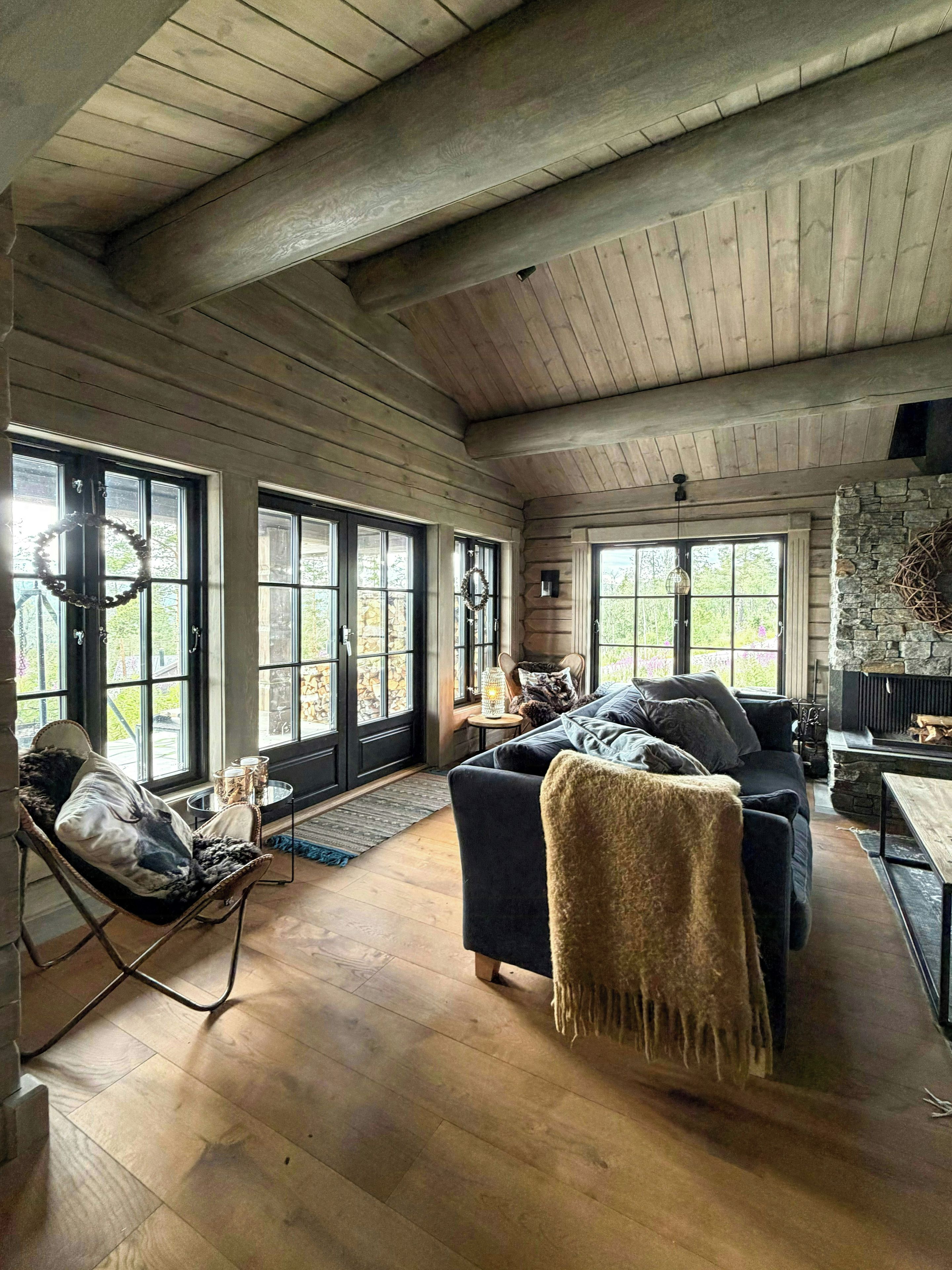 A spacious living room with a high ceiling, wooden beams, a stone fireplace, and large windows.