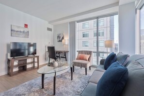 A modern living room with a glass coffee table, a sofa, a television, and a large window offering a city view. - Modern Apt Near Kendall Square (Cambridge, MA)