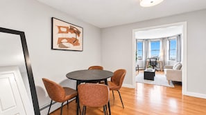 A dining area with a round table and four chairs, a framed abstract painting on the wall, and a view of a living room with a sofa and a desk. - Private Apt Walk to Shops/Restaurants (Boston, MA)