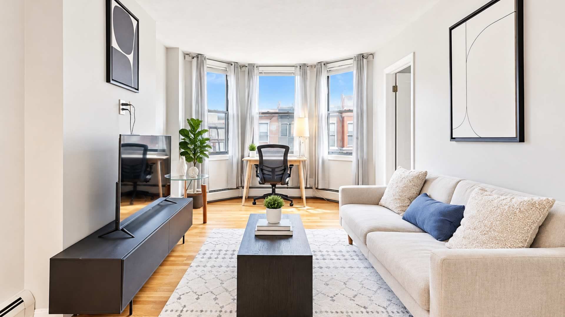 A modern living room with a flat-screen TV, a sofa, a coffee table, and a dining area with a chair and a plant.