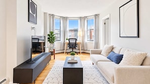 A modern living room with a flat-screen TV, a sofa, a coffee table, and a dining area with a chair and a plant. - Private Apt Walk to Shops/Restaurants (Boston, MA)