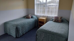 Quarto com duas camas de solteiro, uma pequena mesa de cabeceira de madeira e janela com vista para edifícios.