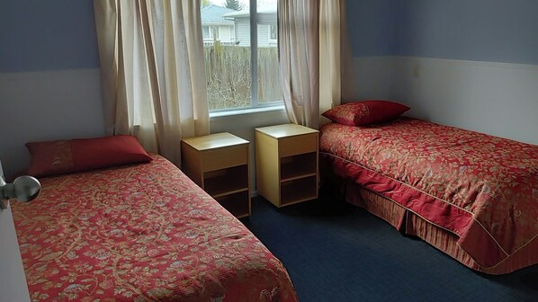 Quarto com duas camas de solteiro, uma pequena mesa de cabeceira de madeira e uma janela com cortinas.