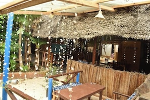 Een rustieke buitenruimte met een rieten dak, houten meubilair en hangende decoraties.