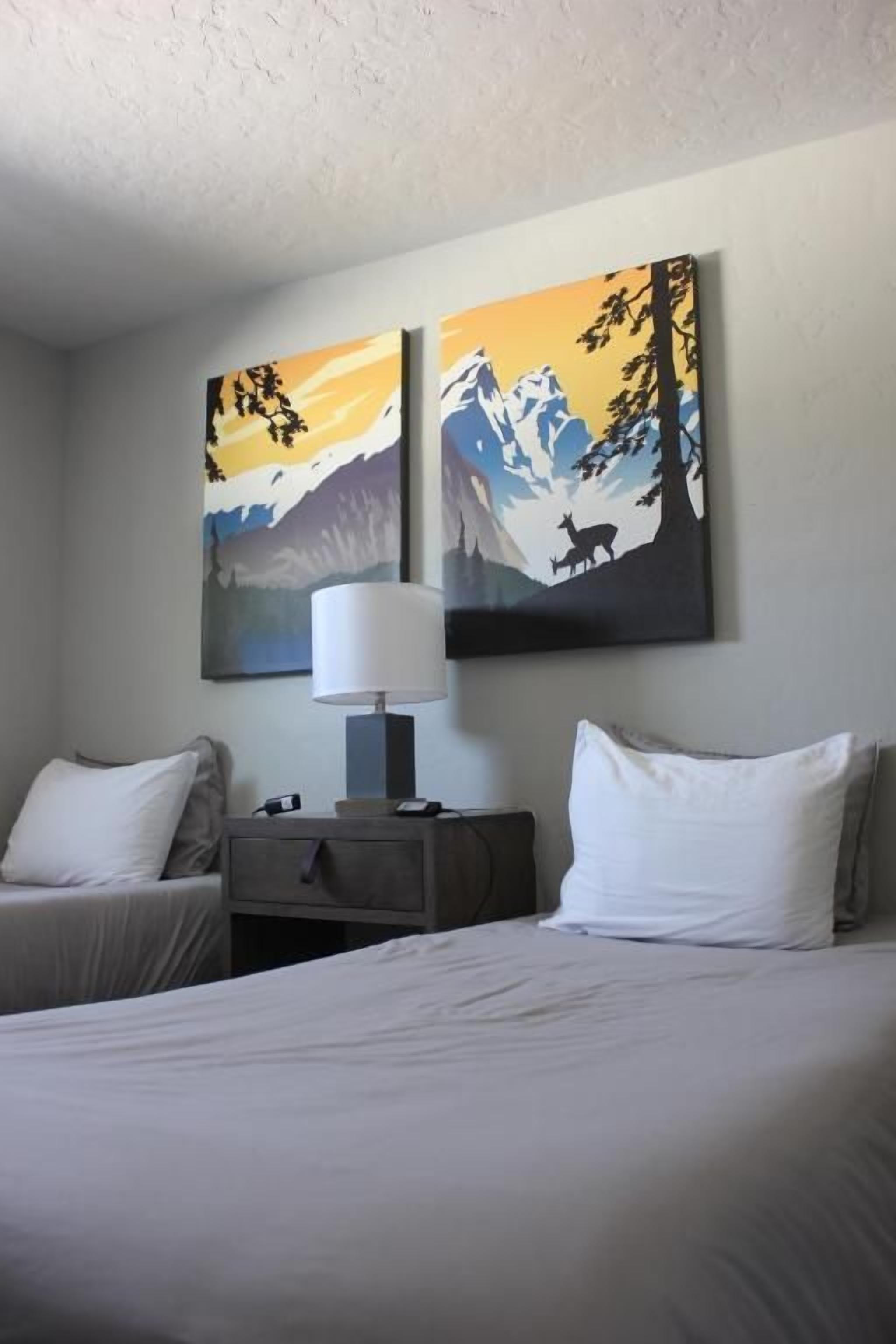 A hotel room with a bed, two bedside tables, a lamp, and two paintings of mountains and wildlife.